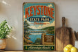 A vintage-style sign for Keystone State Park in Oklahoma, featuring a scenic lake and mountains, is displayed on a shelf next to a wooden cutting board and some lemons.
