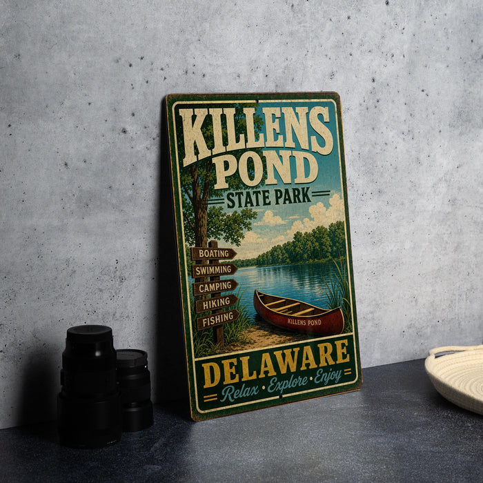 A vintage metal sign for Killens Pond State Park in Delaware, featuring a canoe and surrounded by trees, is displayed on a gray countertop next to a stack of black camera lenses.