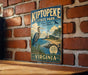 A vintage-style metal sign for Kiptopek State Park, featuring a blue heron and a lighthouse, is displayed on a brick wall.