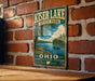 A vintage metal sign for Kiser Lake State Park, featuring a scenic illustration of a lake with boats and trees, mounted on a brick wall.