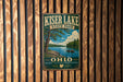 A vintage metal sign for Kiser Lake State Park, featuring a scenic illustration of a lake, trees, and boats, mounted on a wooden wall.