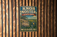A vintage metal sign for Knob Noster State Park in Missouri, featuring a scenic landscape with a cliff and trees.