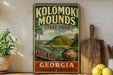 A vintage-style sign for Kolomaki Mounds State Park in Georgia, featuring a scenic landscape with a hill, river, and wooden fence.
