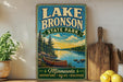 A vintage-style sign for Lake Bronson State Park in Minnesota, featuring a canoe on a lake surrounded by pine trees and mountains.