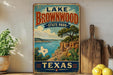 A vintage-style sign for Lake Brownwood State Park in Texas, featuring a scenic landscape with a lake, mountains, and a cactus.