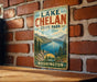 A vintage-style metal sign for Lake Chelan State Park in Washington, featuring a scenic mountain landscape and a lake.