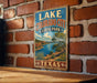 A vintage-style metal sign displaying the text "LAKES COLORADO CITY STATE PARK TEXAS" against a brick wall background.
