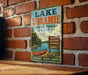 A vintage-style metal sign for Lake Loramie State Park in Ohio, featuring a scenic illustration of a lake, canoe, and surrounding nature.