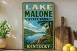 A vintage-style sign for Lake Malone State Park in Kentucky, featuring a scenic landscape with a lake, mountains, and trees.