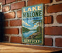 A vintage-style sign for Lake Malone State Park in Kentucky, featuring a scenic landscape with a lake, mountains, and trees.
