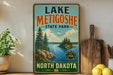 A vintage-style sign for Lake Metigoshe State Park in North Dakota, featuring a scenic landscape with pine trees, a lake, and mountains.