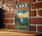 A vintage metal sign for Lake Ouachita State Park in Arkansas, featuring a scenic landscape with pine trees, a lake, and mountains.