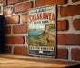 A vintage metal sign for Lake Sakakawea State Park in North Dakota, featuring a scenic illustration of a mountain landscape with a river and a bighorn sheep.