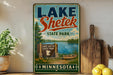 A vintage-style sign for Lake Shetek State Park in Minnesota, featuring a scenic illustration of the park's lake, mountains, and boats.