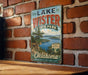A vintage-style sign for Lake Wister State Park in Oklahoma, featuring a scenic landscape with a lake and mountains.