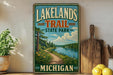 A vintage-style sign for Lakelands Trail State Park in Michigan, featuring a scenic landscape with a river, trees, and a path leading through the greenery.