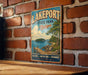 A vintage-style metal sign for Lakeport State Park in Michigan, featuring a scenic landscape with a wooden dock, trees, and a lake.