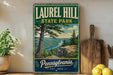 A vintage-style metal sign for Laurel Hill State Park in Pennsylvania, featuring a scenic landscape with a river and pine trees.
