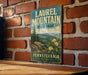 A vintage-style metal sign for Laurel Mountain State Park in Pennsylvania, featuring a scenic landscape with mountains, trees, and flowers.