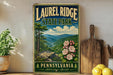 A vintage-style metal sign for Laurel Ridge State Park in Pennsylvania, featuring a scenic mountain landscape with trees, flowers, and a river.