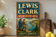 A vintage-style metal sign for the Lewis and Clark Recreation Area in South Dakota, featuring a scenic illustration of two men standing on a hill overlooking a river.
