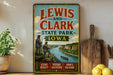A vintage-style sign for Lewis and Clark State Park in Iowa, featuring illustrations of two men on a cliff overlooking a river and a boat.