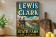 A vintage-style sign for Lewis and Clark State Park in North Dakota, featuring a scenic illustration of two hikers on a trail overlooking a river.