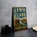 A vintage-style metal sign for Lewis and Clark State Park in North Dakota, featuring an illustration of two hikers on a trail overlooking a river.