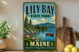 A vintage-style metal sign for "Lily Bay State Park" in Maine, featuring a scenic landscape with a lake, mountains, and a canoe.