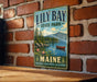 A vintage-style metal sign for "Lily Bay State Park" in Maine, featuring a scenic landscape with a lake, mountains, and a canoe.