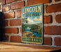 A vintage metal sign for Lincoln State Park in Indiana, featuring a scenic landscape with a river, trees, and a building.