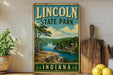 A vintage-style sign for Lincol State Park in Indiana, featuring a scenic landscape with a lake, mountains, and a cabin.
