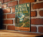 A vintage-style metal sign for the Lincoln Trail in Illinois, featuring a scenic illustration of a path leading through a lush, wooded landscape with a mountain backdrop.