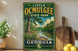 A vintage-style sign for Little Ocmulgee State Park in Georgia, featuring a scenic landscape with a river and trees.