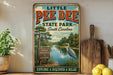 A vintage-style metal sign for Little Pee Dee State Park in South Carolina, featuring a scenic image of a lake, trees, and a bird.
