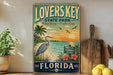 A vintage-style sign for "Lovers Key State Park" in Florida, featuring a beach scene with a heron, palm trees, and a sunset.