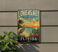 A vintage-style sign for "Lovers Key State Park" in Florida, featuring a beach scene with palm trees, a crane, and a sunset.