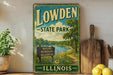 A vintage-style sign for Lowden State Park in Illinois, featuring a scenic landscape with a river, trees, and a sign indicating the park's nature, recreation, relaxation, and nature.