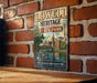 A vintage metal sign for Lowell Heritage State Park, featuring an illustration of a bridge and buildings, mounted on a brick wall.