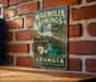 A vintage-style metal sign for Magnolia Springs State Park in Georgia, featuring a scenic image of the park and its surroundings.