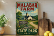A vintage-style sign for Malabar Farm, featuring a farm scene with a barn, trees, and a dirt road, is displayed on a shelf next to a wooden cutting board and some lemons.