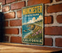 A vintage-style metal sign for Manchester State Park in California, featuring a coastal landscape with cliffs, flowers, and the ocean.