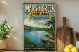 A vintage-style metal sign for Marsh Creek State Park in Pennsylvania, featuring a scenic illustration of a lake, dock, and sailboat.