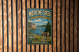 A metal sign with a scenic image of a deer in a wooded area, with a lake and mountains in the background, mounted on a wooden wall.