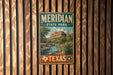 A vintage metal sign for Meridian State Park in Texas, featuring a scenic landscape with mountains, a river, and desert vegetation.