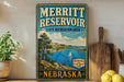 A vintage-style sign for the Merritt Reservoir State Recreation Area in Nebraska, featuring a scenic landscape with a lake, mountains, and a sailboat.