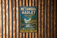 A vintage metal sign for the Metamora-Hadley Recreation Area in Michigan, featuring a scenic landscape with a waterfall and pine trees.