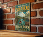 A vintage-style metal sign for Mike Roess Gold Head Branch State Park in Florida, featuring a scenic illustration of a river with people kayaking.
