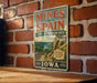 A vintage-style sign for the Mines of Spain State Recreation Area in Iowa, featuring a scenic illustration of a mountain landscape with a river and a wooden bridge.