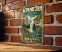 A vintage-style metal sign for Minnesota State Park hangs on a brick wall, depicting a scenic waterfall surrounded by lush greenery.